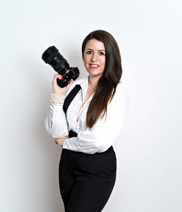 A black-and-white photo of Diana Kiss, a photographer in Chicago, leaning against a tree with her camera raised, wearing a black sheer top and black pants, and smiling for a headshot photo