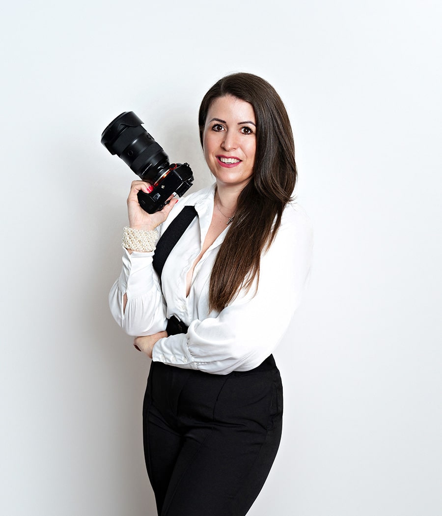 A black-and-white photo of Diana Kiss, a photographer in Chicago, leaning against a tree with her camera raised, wearing a black sheer top and black pants, and smiling for a headshot photo